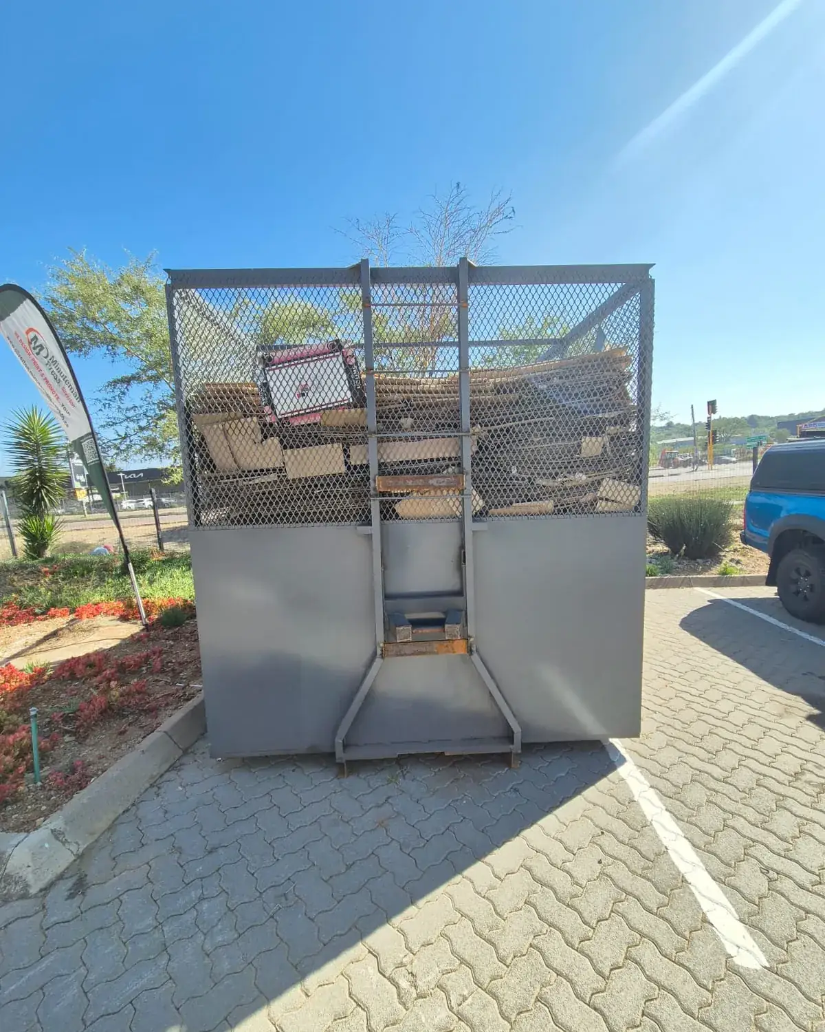 A Murero mesh cage standing in a retail complex parking bay, loaded with flattened cardboard.
